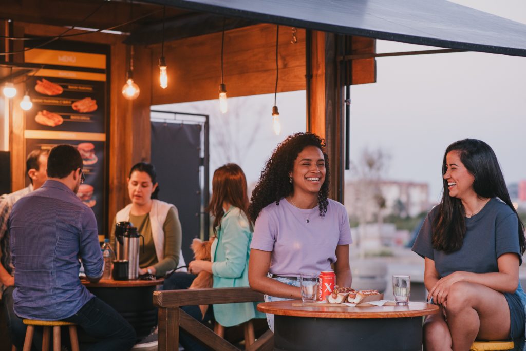 Dois amigos sorrindo e conversando em um restaurante ao ar livre, com luzes pendentes iluminando o ambiente.