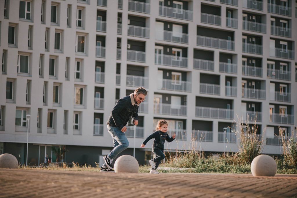 Pai e filho correndo juntos em um parque, com um prédio moderno ao fundo.