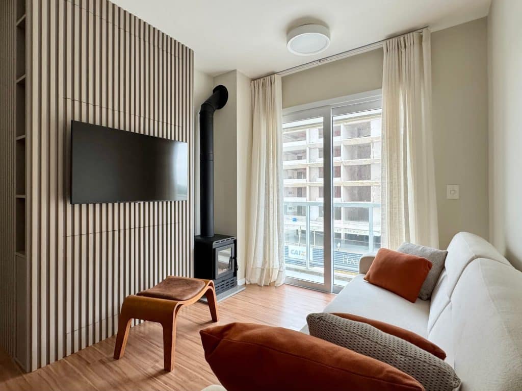 Sala de estar moderna e minimalista com parede de madeira listrada, TV montada na parede e um aquecedor a lenha. O ambiente conta com um sofá claro e almofadas coloridas, além de uma janela com cortinas claras, oferecendo vista para uma obra em construção.