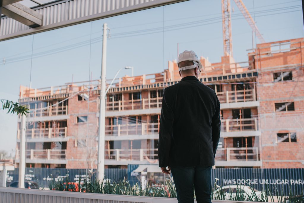 Homem de capacete observa obra de prédio em construção com estrutura de tijolos e andaimes expostos.