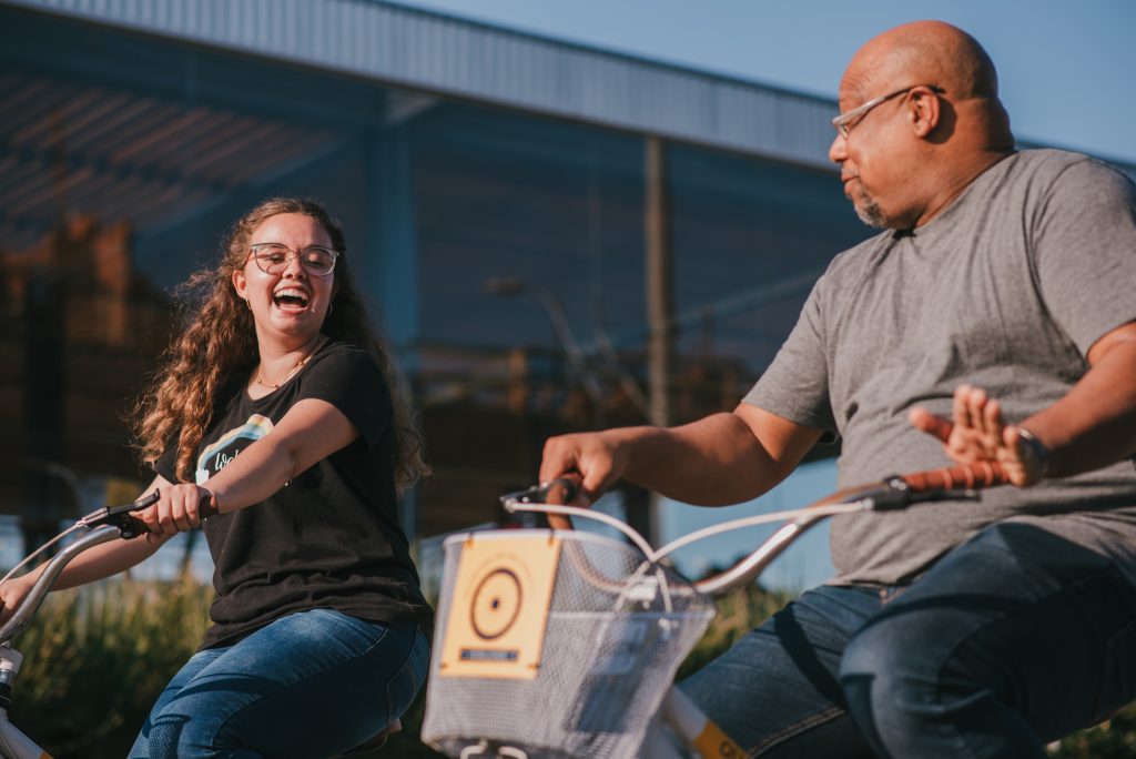 Mulher e homem andam de bicicleta sorrindo um para o outro em área urbana com sol forte.
