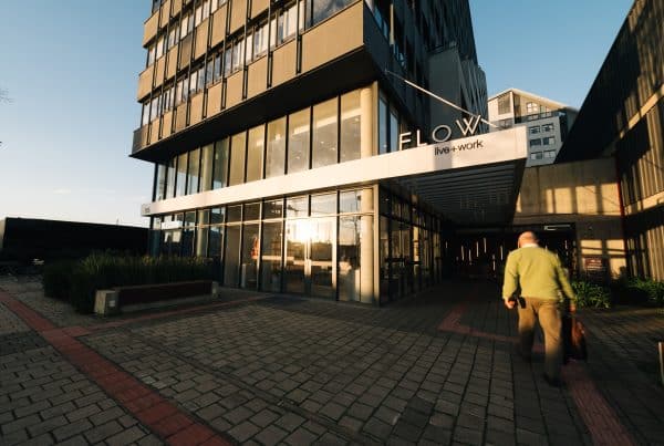 Fachada de um prédio moderno com grandes janelas de vidro refletindo a luz do sol, uma placa com os dizeres 'FLOW live + work' e uma pessoa caminhando em direção à entrada, carregando uma bolsa.