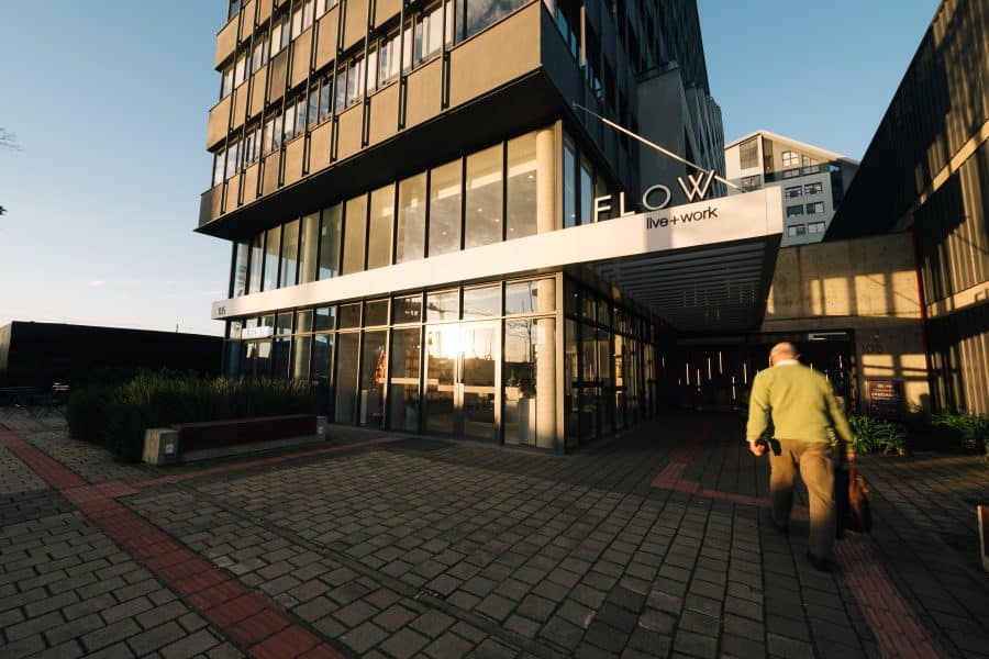 Fachada de um prédio moderno com grandes janelas de vidro refletindo a luz do sol, uma placa com os dizeres 'FLOW live + work' e uma pessoa caminhando em direção à entrada, carregando uma bolsa.