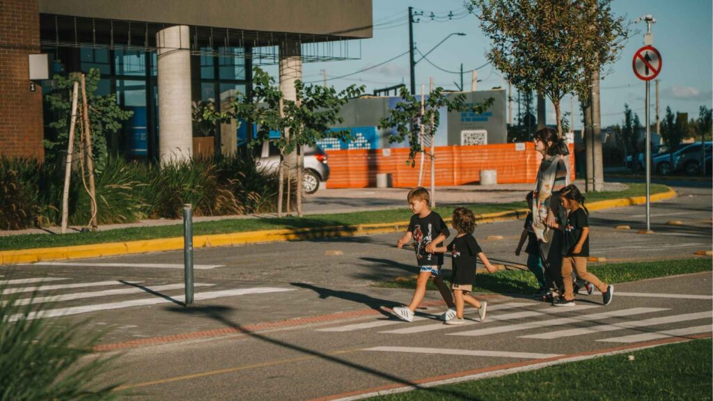 Mulher acompanhada de crianças atravessando a faixa de pedestres em rua urbana com sinalização viária, árvores jovens, prédios modernos e área em obras ao fundo, retrato da mobilidade urbana e do dia a dia familiar em bairros residenciais de Pelotas.