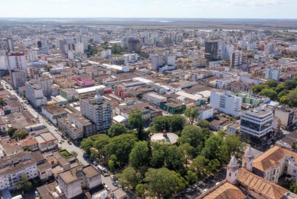 Vista aérea do centro urbano com prédios de diferentes alturas, quadras bem definidas, áreas arborizadas e igreja histórica em destaque, panorama que mostra a diversidade arquitetônica, comercial e residencial dos bairros de Pelotas.