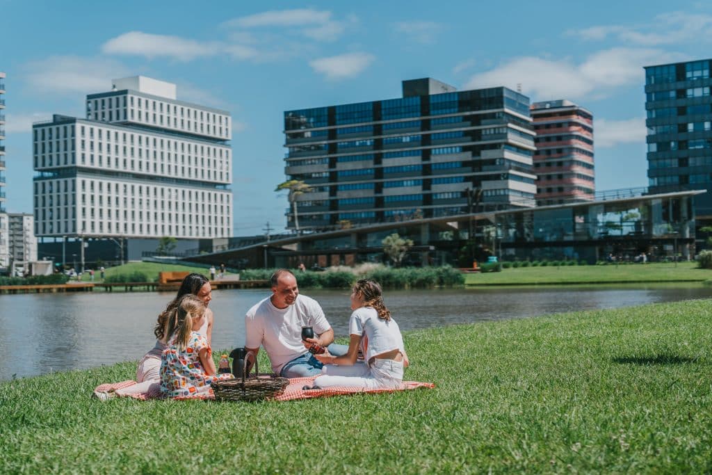 A imagem mostra uma família desfrutando de um piquenique em um parque ao lado de um lago. No primeiro plano, há um homem, uma mulher e duas crianças sentadas sobre uma toalha quadriculada. Eles estão sorrindo e interagindo, com uma cesta de piquenique ao lado. Ao fundo, é possível ver edifícios modernos, com um ambiente urbano bem desenvolvido e espaços verdes ao redor. O céu está limpo e o dia é ensolarado, transmitindo uma sensação de relaxamento e lazer.