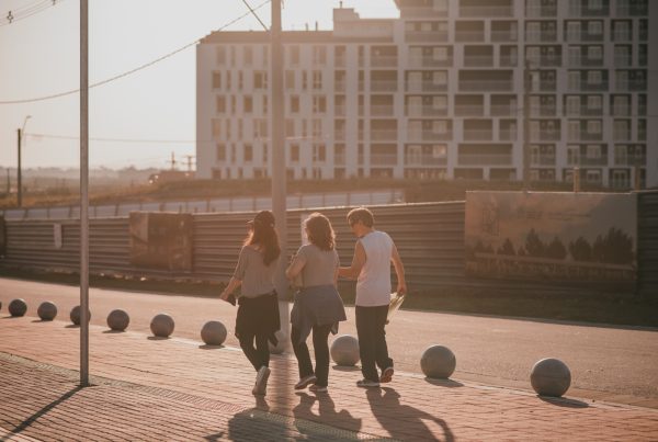 Três pessoas caminhando juntas ao entardecer, com luz suave e um prédio residencial ao fundo.
