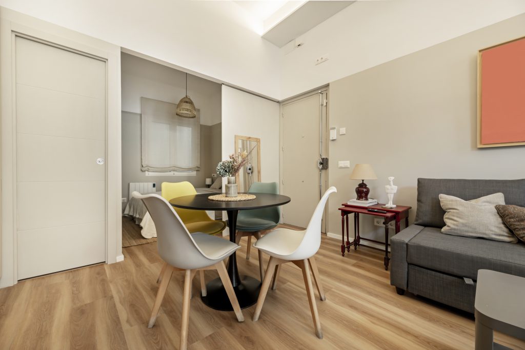 Sala compacta com piso de madeira clara, mesa de jantar redonda com cadeiras coloridas e uma decoração simples, com uma parede de tom neutro ao fundo.