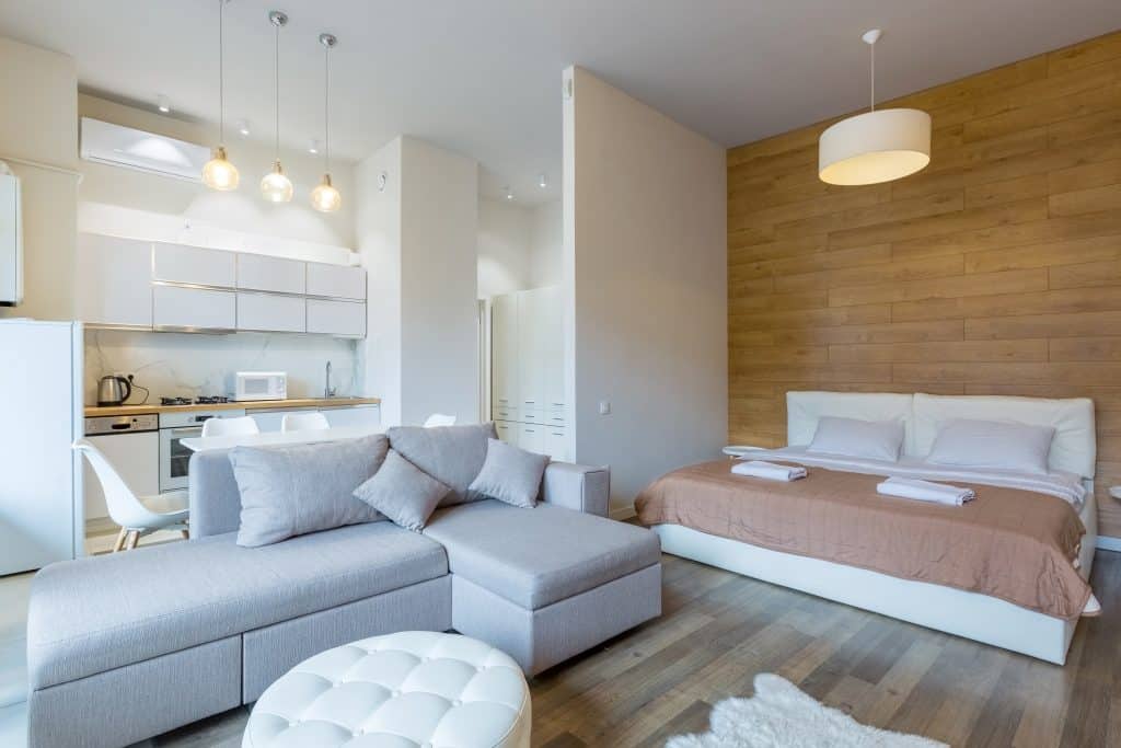 Interior de um apartamento moderno e minimalista, com sala de estar integrada à cozinha e um quarto com cama de casal. O ambiente tem móveis neutros, piso de madeira e uma parede de madeira atrás da cama.
