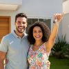Casal sorridente em frente à casa nova, com a mulher segurando chaves no alto em gesto de celebração.