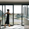 Mulher de vestido preto abrindo uma porta de vidro deslizante em apartamento com vista panorâmica para lago, parque e prédios modernos.