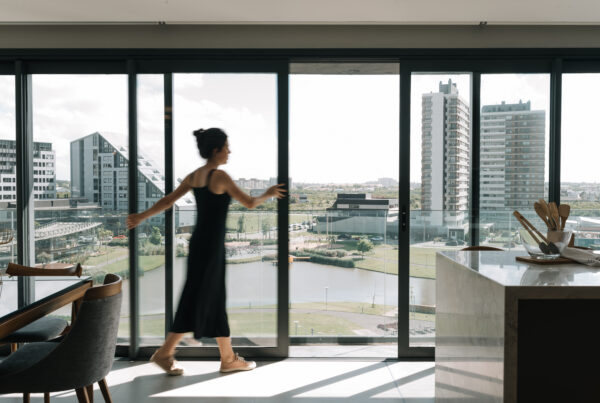 Mulher de vestido preto abrindo uma porta de vidro deslizante em apartamento com vista panorâmica para lago, parque e prédios modernos.