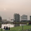 Vista panorâmica de um parque urbano moderno em Pelotas ao entardecer, com um lago artificial no centro e pessoas sentadas no gramado. Ao fundo, destacam-se edifícios residenciais altos e contemporâneos de um bairro planejado.