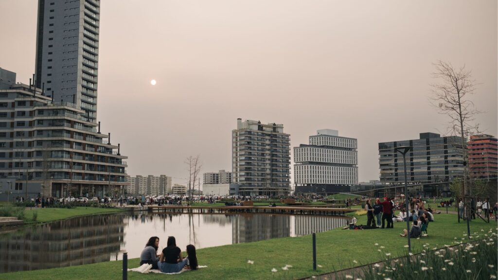 Vista panorâmica de um parque urbano moderno em Pelotas ao entardecer, com um lago artificial no centro e pessoas sentadas no gramado. Ao fundo, destacam-se edifícios residenciais altos e contemporâneos de um bairro planejado.